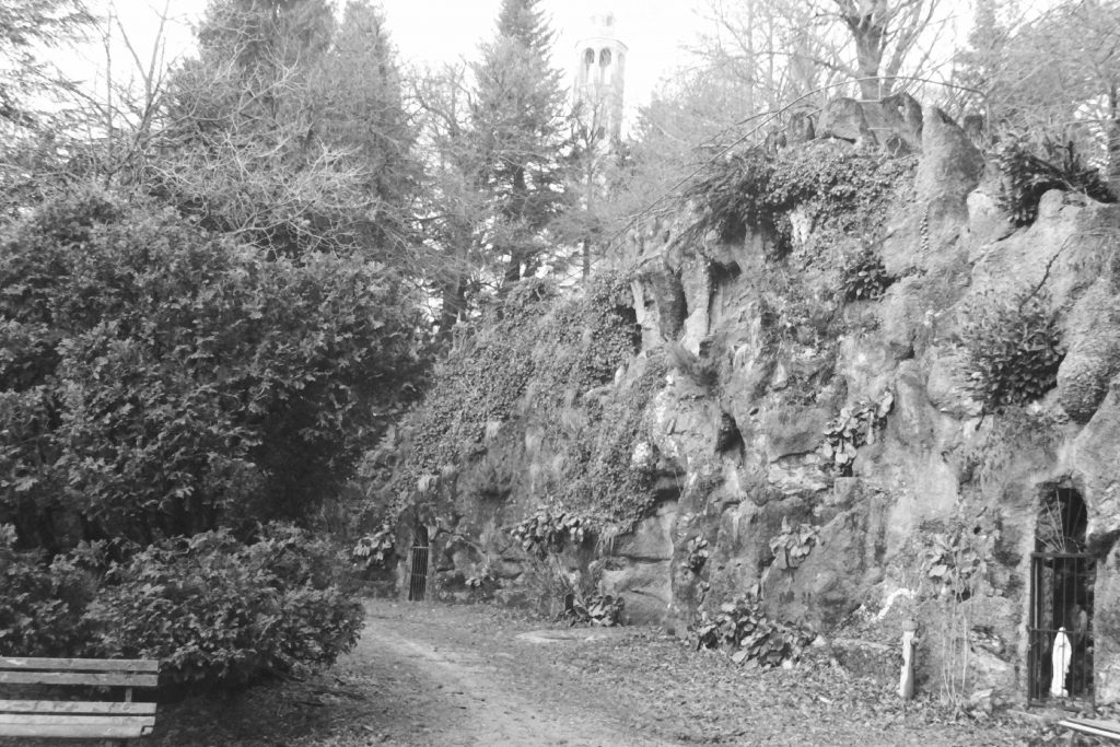 Black and white photograph of a stone hillside with small alcoves set into, behind gates are holy statues