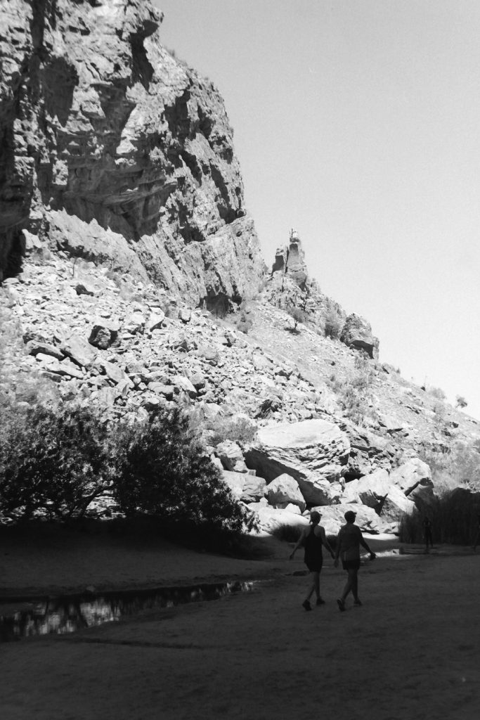 Black and white photograph of two women walk alongside a shallow creek bed beneath the face of a gorge