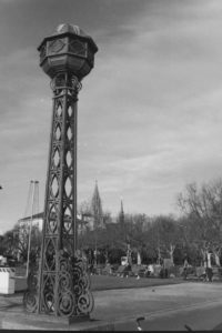 Black and white photograph of a park with a tall iron sculpture in the foreground.