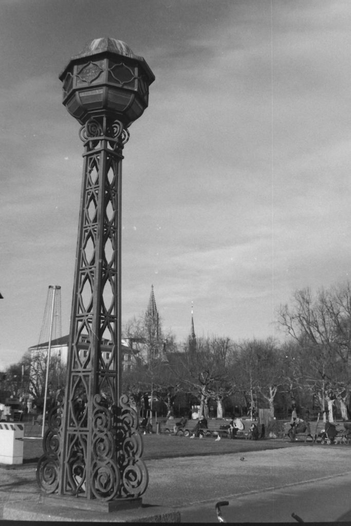 Black and white photograph of a park with a tall iron sculpture in the foreground.