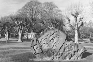 Black and white photograph of a park with bar trees and and a large stone sculpture in the foreground