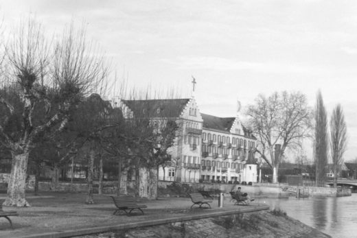 Black and white photograph of a park and buildings along the edge of lake, a man is sitting on park bench