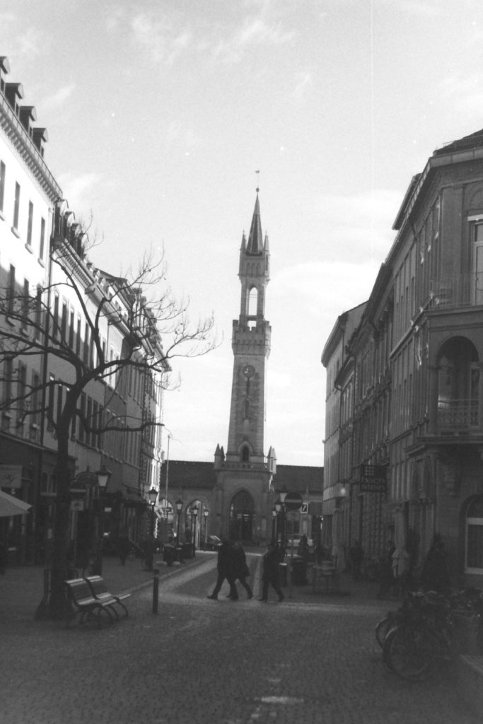 Black and white photograph of a town square with a spire at the far end an people walking through it