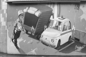 Black and white photograph of a mural painted on a wall. The mural shows a car parked in a disabled parking spot with it's tyres clamped and the word "Karma".