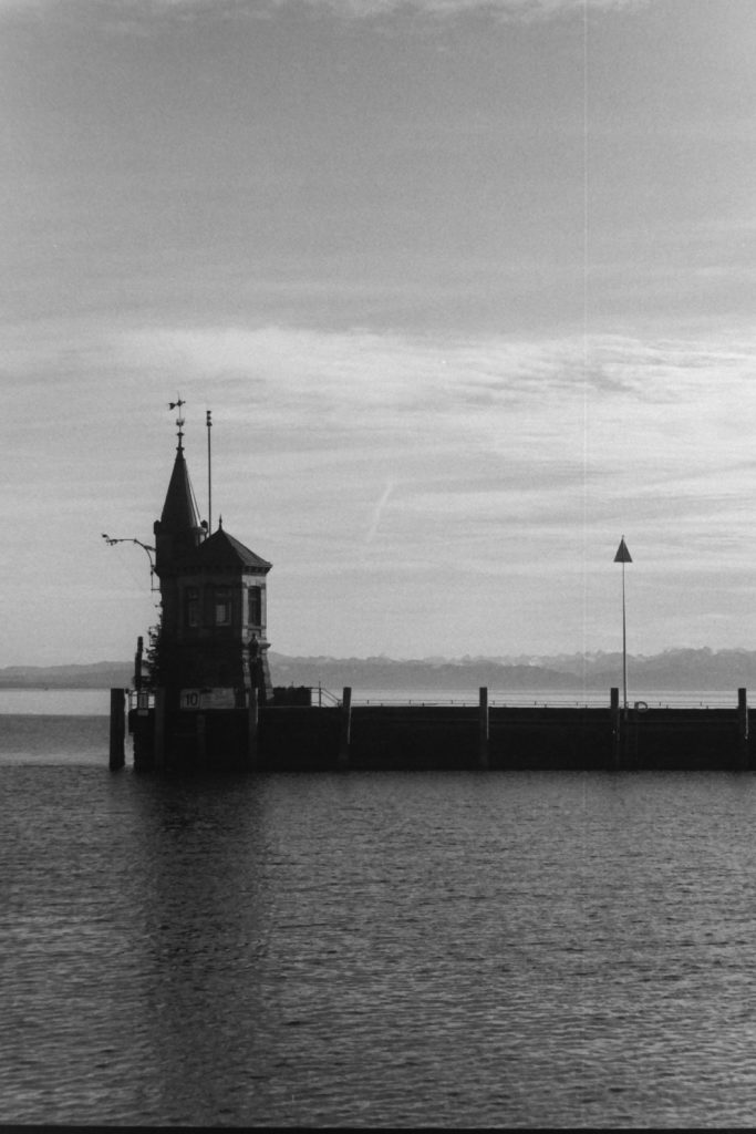 Black and white photograph of a large lake with a jetty in the middle-ground. At the end of the jetty is a small building.