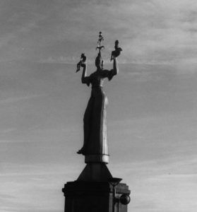 Black and white photograph of a concrete statue of a woman wearing a fools cap on her head and holding her arms up. On her hands sit two small figures.