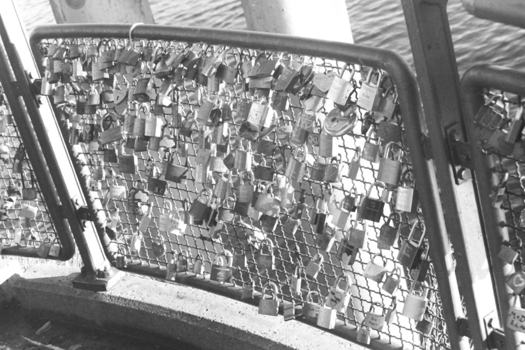 Black and white photograph of a fence covered in love locks (padlocks with names and initials inscribed).