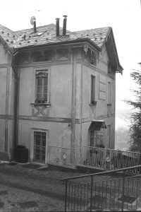 Black and white photograph of a tqo story house on hillside with a paved path and stair case rail in the foreground