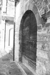 Black and white photograph of a wooden door set into a stone building, there is a large metal slider across the door, and the number 20 to the side