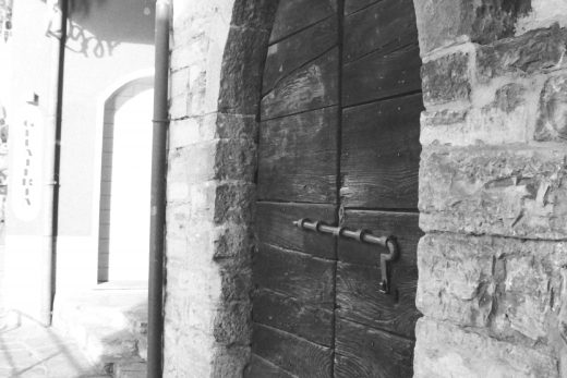 Black and white photograph of a wooden door set into a stone building, there is a large metal slider across the door, and the number 20 to the side