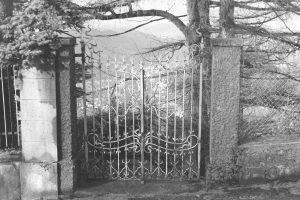 Black and white photograph of a wrought iron gate in a fence with the view of a valley behind it