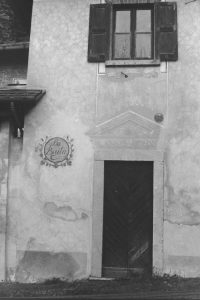 Black and white photograph of the wooden door of a stone building, there is a window above the door with shutters and a an engraving to the side that says "La Baila