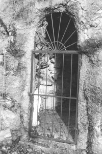 Black and white photograph of an iron gate at the entrance to an alcove, behind the gate is a holy statue