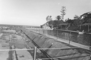 Black and white photograph of swimming pools with the ocean in the background and people sitting around the edges of the pools