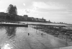 Black and white photograph of a swimming pool with houses and the ocean in the background and two people swimming in the pool