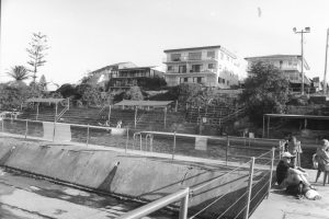 Black and white photograph of swimming pools with houses in the background and people sitting around the edges of the pools