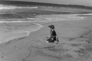 Black and white photograph of a boy sitting on the beach close to the water's edge, looking at the waves coming in