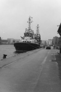 Black and white photograph of a marina with a car driving past a moored tugboat