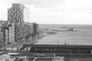 Black and white photograph of a harbour lined with apartment buildings, in the foreground is a train stopped at a station