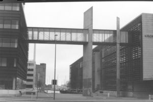 Black and white photograph of a promenade with two large building either side and a high glass walkway between the two