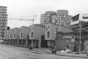 Black and white photograph of a street lined with modern square town houses with two large round apartment buildings behind them