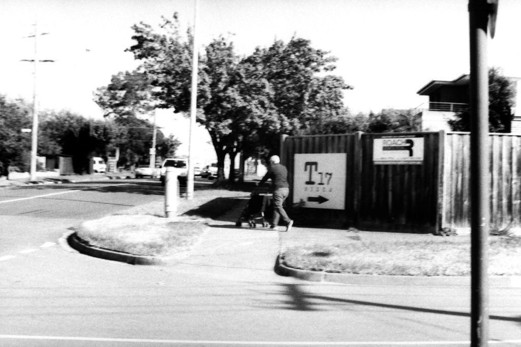 Black and white photograph of a man pushing a pram around the corner of a suburban street