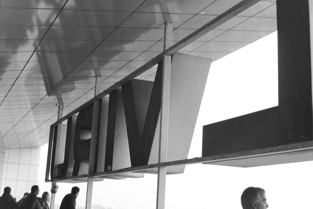 Black and white photograph of people standing on a hotel balcony beneath the hotel sign that reads "VIRU"backwards.