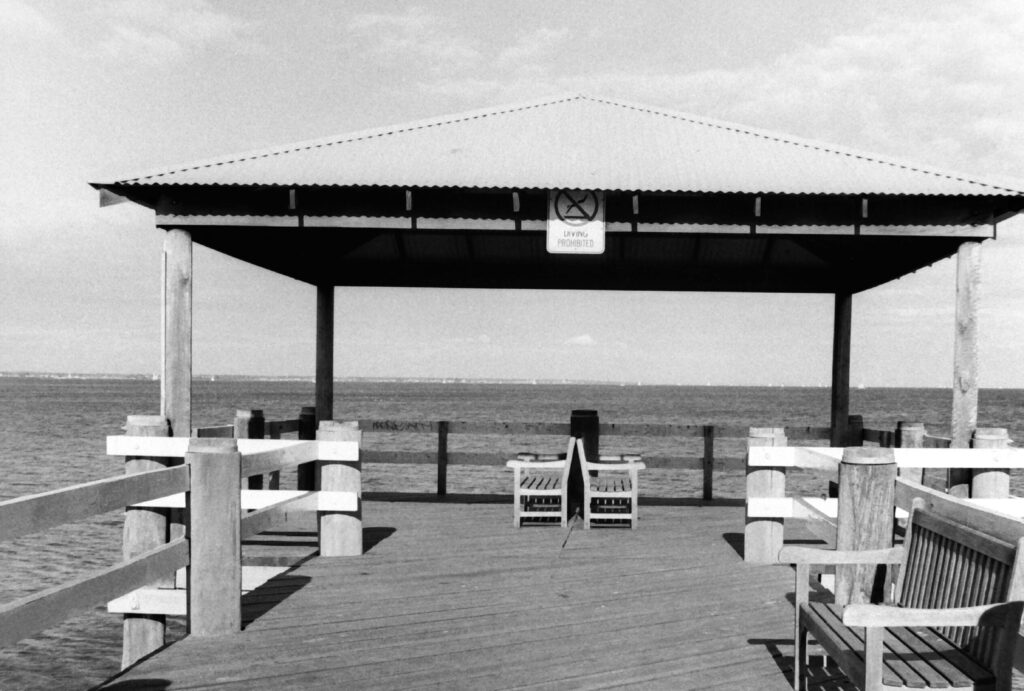 Black and white photo of a pier with bench seats looking out over the water