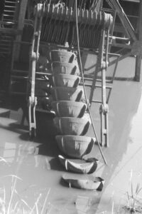 Black and white photo of the buckets of a dredging machine extending into water.