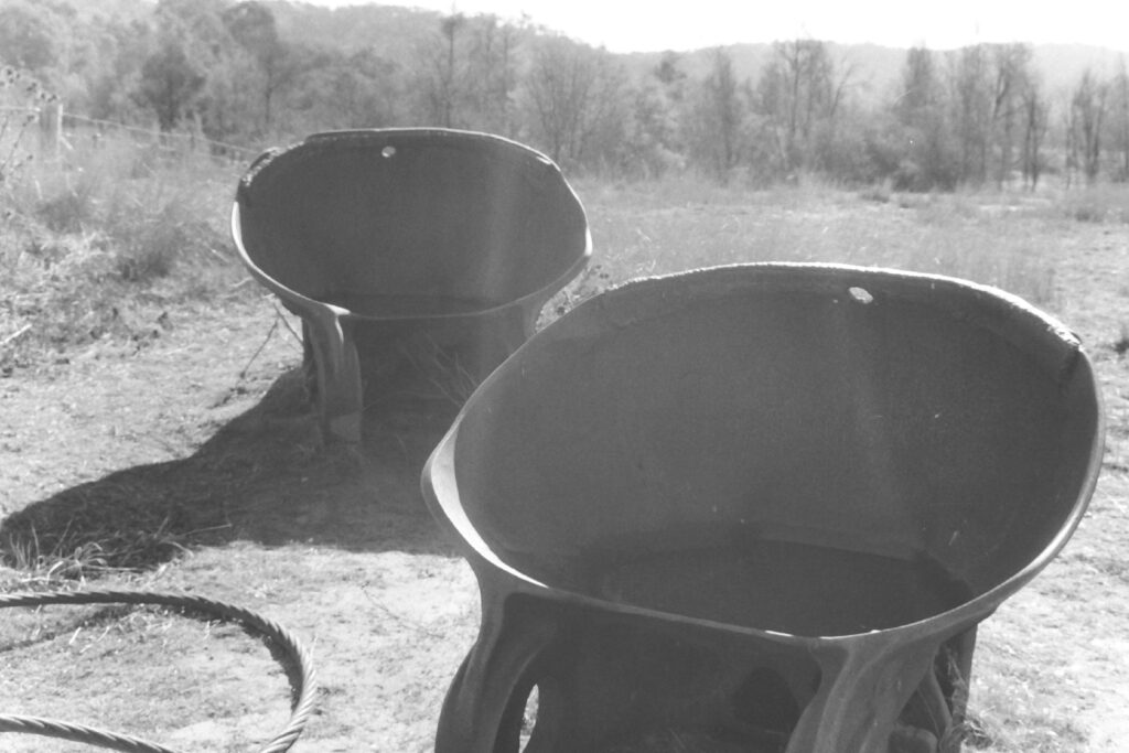 Black and white photograph of two large dredging buckets on land, with a coil of wire rope visible to the side. Bushy grass and trees are in the background.