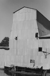 Black and white photograph of a large building clad in corrugated iron, with the water from the dredge visible at the lower edge.