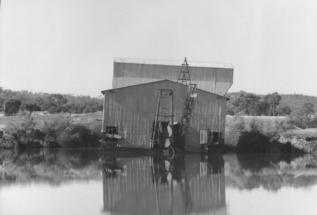 Black and white photograph of a large building with dedging machinery leaning out of a body of water. The dredge is reflected in the water and leaning to the right of the image.