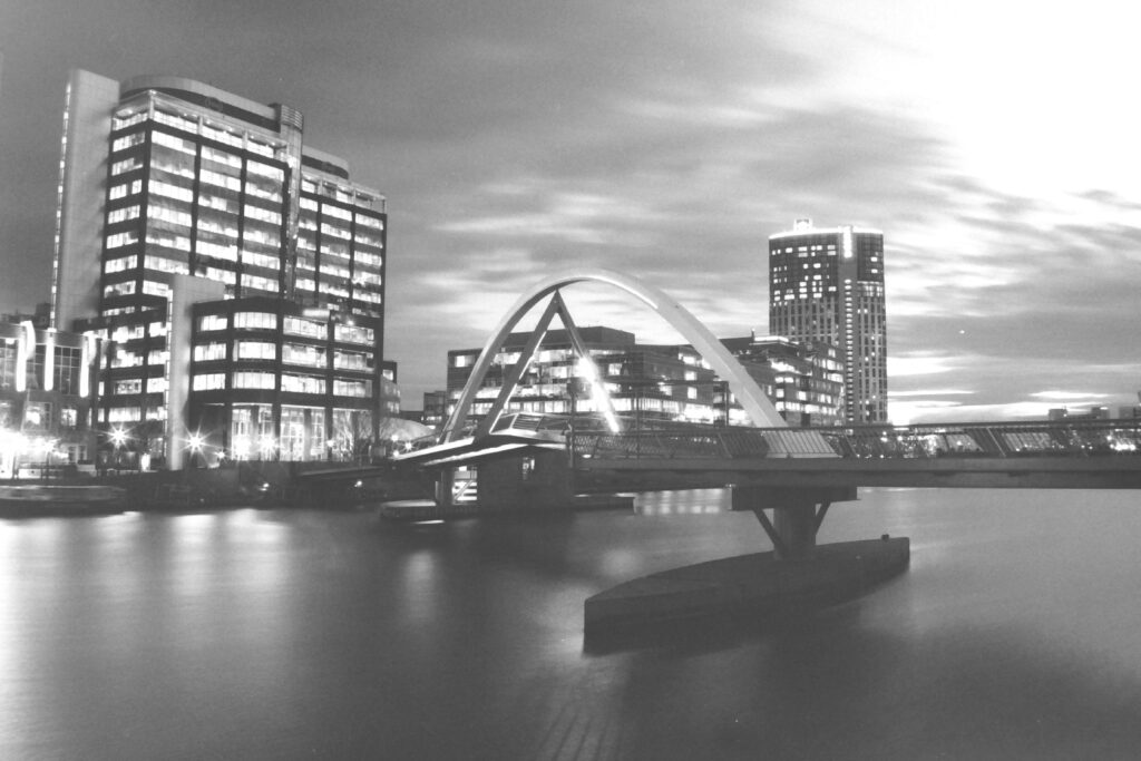 Black and white photo taken at night of a pedestrian bridge spanning a river. The bridge has an arch over the top and is lit up. The surrounding buildings across the water are also lit up.