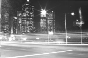 Black and white photo of a city street taken at night. There is a streak of light across the image from passing cars. Skyscrapers are in the background.