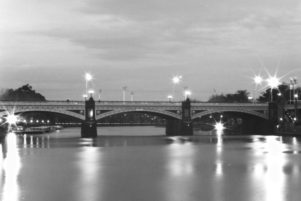 Black and white photo at night of a bridge spanning a river. There are boats moored on one side, and in the background are the lights of an outdoor sports stadium.