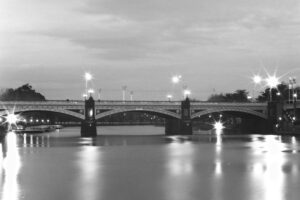 Black and white photo at night of a bridge spanning a river. There are boats moored on one side, and in the background are the lights of an outdoor sports stadium.