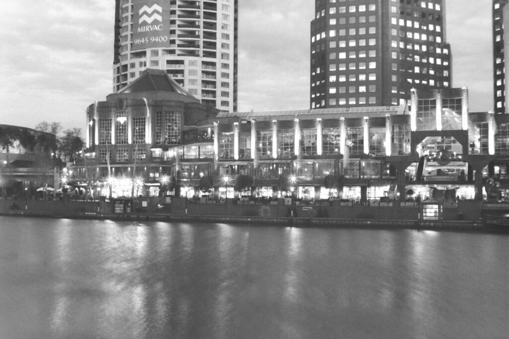 Black and white photo across a river to a promenade of brightly lit buildings and outdoor eating areas.