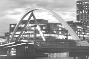 Black and white photo of a pedestrian bridge at night. The bridge has a curved structure over the top of it and angles up into the middle. There are buildings in the background.