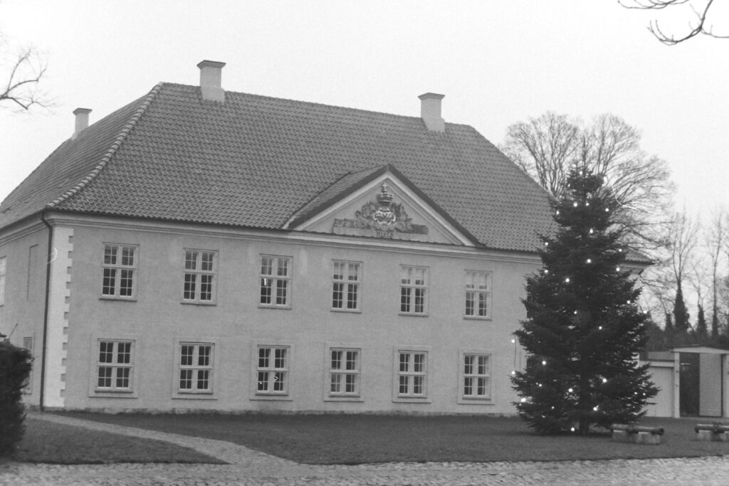 Black and white picture of a large stone building with two levels of windows and a crest above them. There is a tall tree decorated with lights and two cannons on the lawn in front.