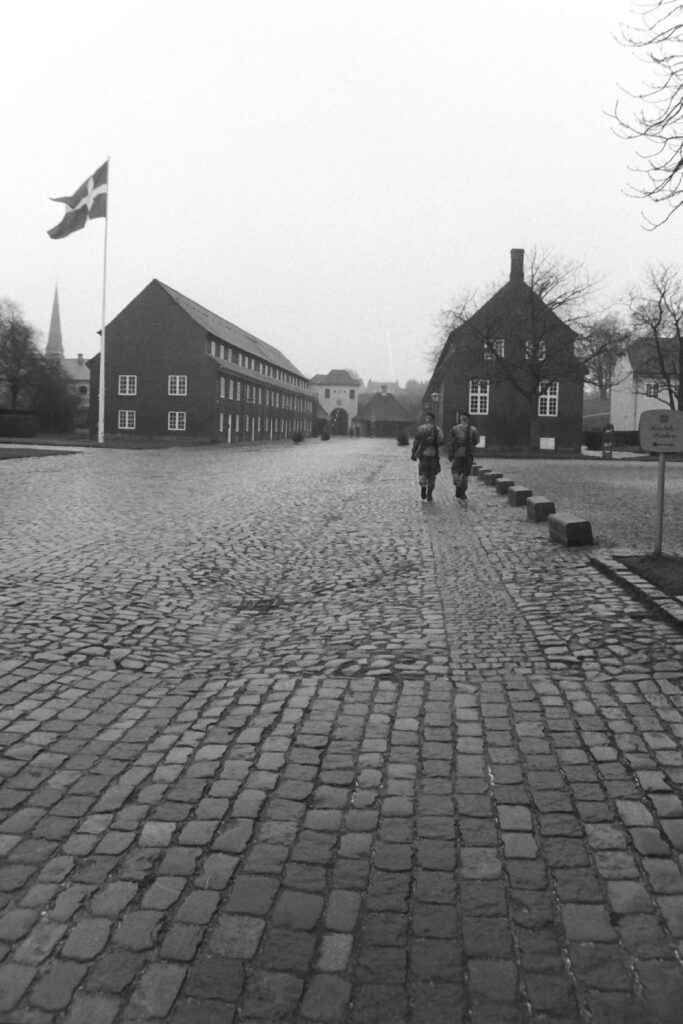 Black and white photo taken looking down a cobbled road leading towards two barracks buildings. Two soldiers are walking towards the barracks and there is a flagpole with the Danish flag.