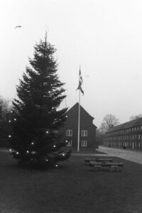 Black and white photograph of a lawn with a large tree decorated with lights and a star on top, a flagpole flying the Danish flag and two canons. In the background is paved roads and barracks buildings.