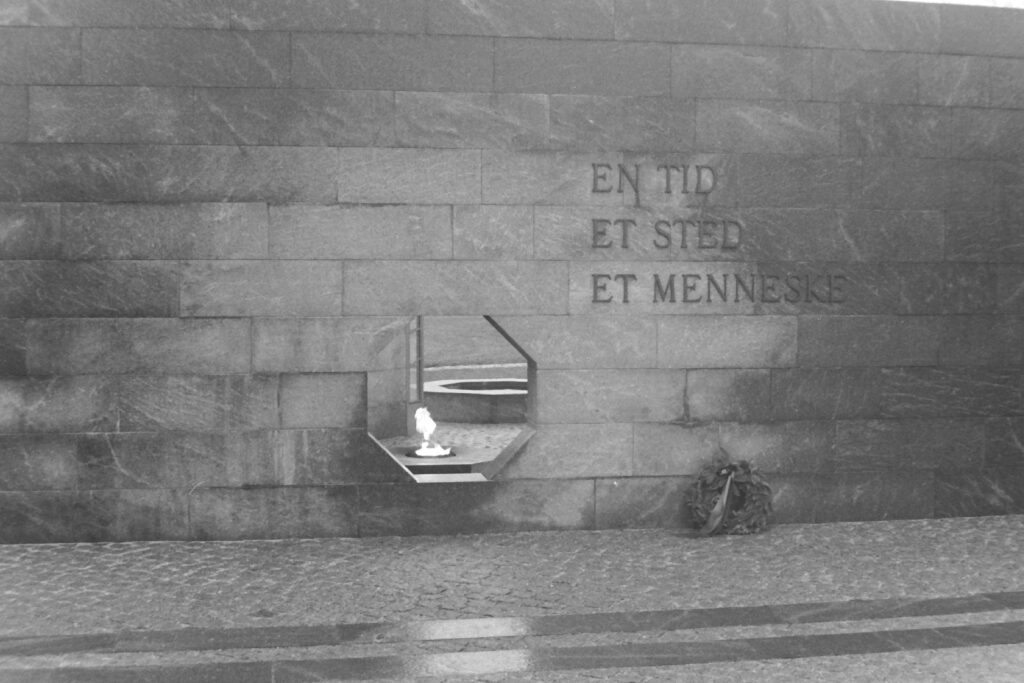 Black and white photo of a large stone wall with an eternal flame and a wreath laid next to it. The inscription on the wall reads "EN TID ET STED ET MENNESKE".