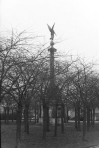 Black and white photo of a tall monument surrounded by trees, at the top, rising out of the trees is an angel.