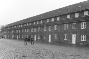 Black and white photo of a long barracks building with two soldiers walking past on a cobbled road.