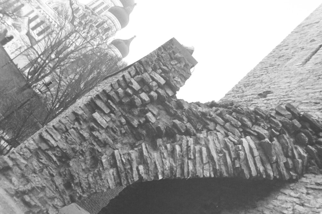 Black and white photo, taken on an angle, with a stone wall and archway in the foreground and an church with onion domes in the background.