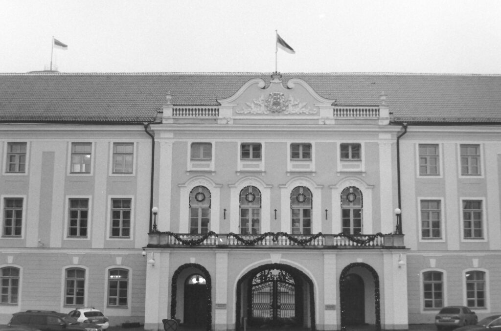 Black and white photo of a grand stone building with a balcony and coach entrance. the flag of Estonia is flying above it.