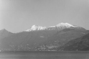 Black and white photo of a mountain range with snow on the peaks and a large village and lake in the foreground.