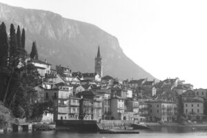 Black and white photo of a lake side village with a clock tower and mountains in the background.