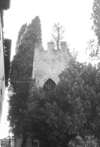 Black and white photo of a stone medieval tower with turrets mostly hidden by trees.
