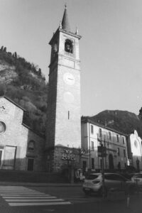 Black and white photo of a stone church with a belltower and clock. In the background are mountains, and in the foreground is a street with pedestrian crossing and cars.